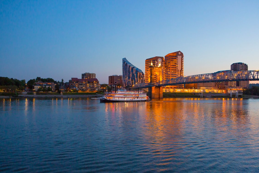Covington-Kentucky | Rollins Insurance Riverfront cityscape at dusk with illuminated buildings and a blue bridge reflecting on the river