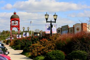 Red clock tower in a shopping plaza with decorative lampposts, banners, and storefronts along a sunny sidewalk lined with bushes and flowers.
