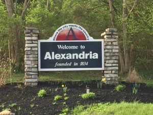 Welcome sign for Alexandria with stone pillars, reading 'Welcome to Alexandria, founded in 1834' amid greenery.