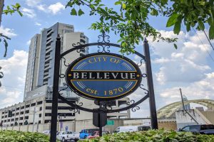 Bellevue city sign: blue and gold crest reading 'City of Bellevue, est. 1870' on an ornate black frame, with modern buildings behind.