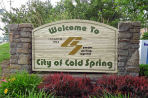 Wooden welcome sign for City of Cold Spring, flanked by stone pillars and landscaped greenery.