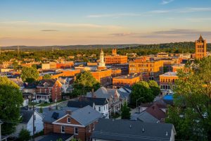 Aerial view of a small city at sunset with red brick buildings, tree-lined streets, and a tall church-like tower on the right.