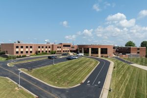 Brick school building with curved driveway and parking lot under a blue sky; manicured lawn surrounds the campus.