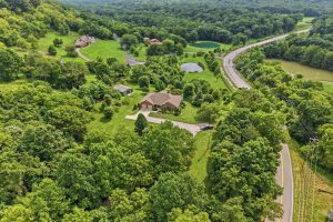 Aerial view of a brick house with a long driveway, large green yard, and dense trees surrounding the property near a winding road and ponds nearby.