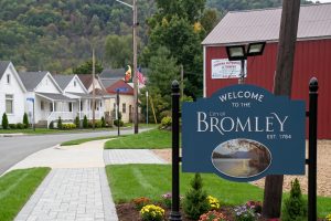 Blue 'Welcome to the City of Bromley, Est. 1784' sign beside a paved sidewalk, with colorful flowers and a residential street or hillside in the background.