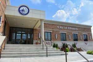 Exterior view of the City of Elsmere government building with brick facade and steps leading to glass doors, festive bunting, and a circular city seal at the top left.