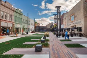 Pedestrian shopping street with pastel storefronts, outdoor café seating, and a brick-paved central walkway under a blue sky.