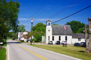 White church with a small bell tower on a sunny street, American flag flying in front and cars parked along the curb, trees and power lines in the background.