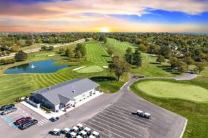 Aerial view of a golf course at sunset, showing green fairways, a water hazard, trees, and a clubhouse with parking in the foreground.