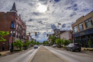 Downtown street scene with brick storefronts, trees, and cars under a cloudy sky with an American flag above.