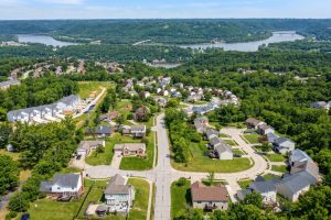 Aerial view of a suburban neighborhood with curved streets, green lawns, and rows of houses, hills and a river with bridges in the distance