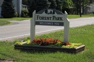 Forest Park welcome sign mounted on white posts beside a flower bed along a street.